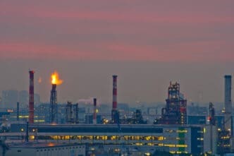 A Russian oil refinery with multiple smokestacks and a large flare stack burning against a hazy, pink-and-purple sunset sky