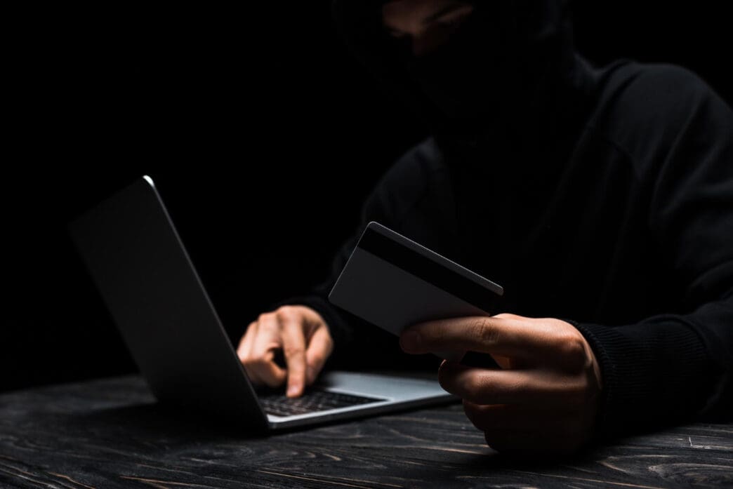 A person in a black mask and hoodie holds a credit card while typing on a laptop