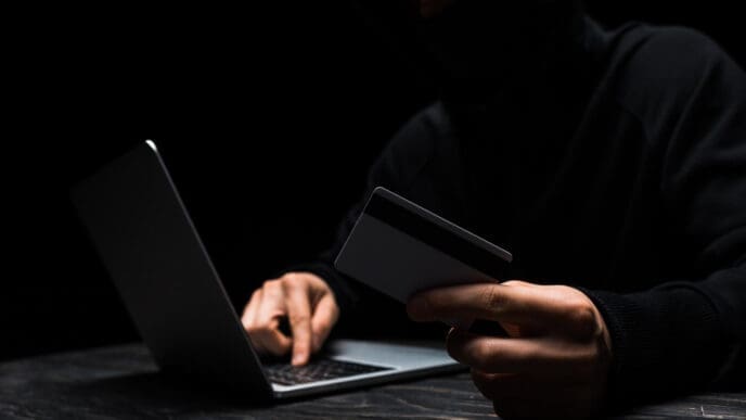 A person in a black mask and hoodie holds a credit card while typing on a laptop