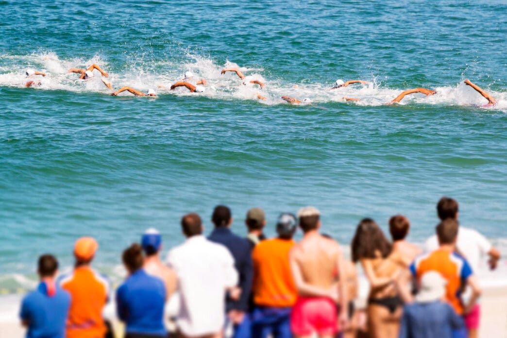 Swimmers in the ocean compete in an open-water race as spectators watch from a beach.