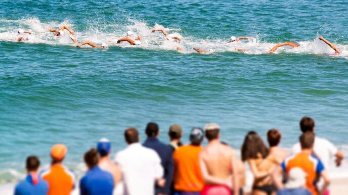 Swimmers in the ocean compete in an open-water race as spectators watch from a beach.