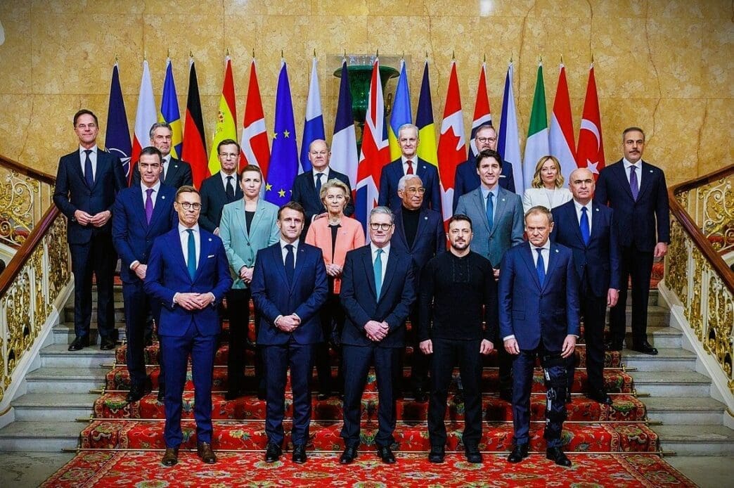 A group of world leaders, including President Volodymyr Zelenskyy, are posed for a group photo on a grand staircase with many international flags behind them.