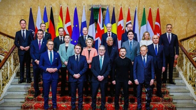 A group of world leaders, including President Volodymyr Zelenskyy, are posed for a group photo on a grand staircase with many international flags behind them.