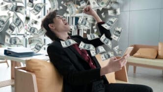 An excited man in a suit looks at his smartphone as dollar bills rain down around him, symbolizing financial success.