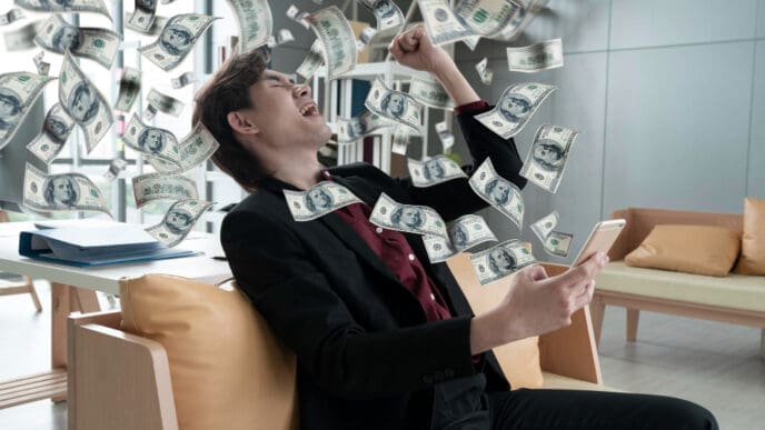 An excited man in a suit looks at his smartphone as dollar bills rain down around him, symbolizing financial success.