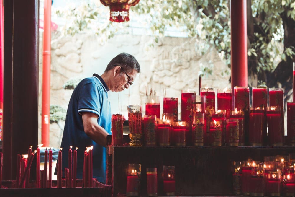 An elderly man in a blue shirt standing in a Chinese temple, paying tribute in front of many lit red candles.