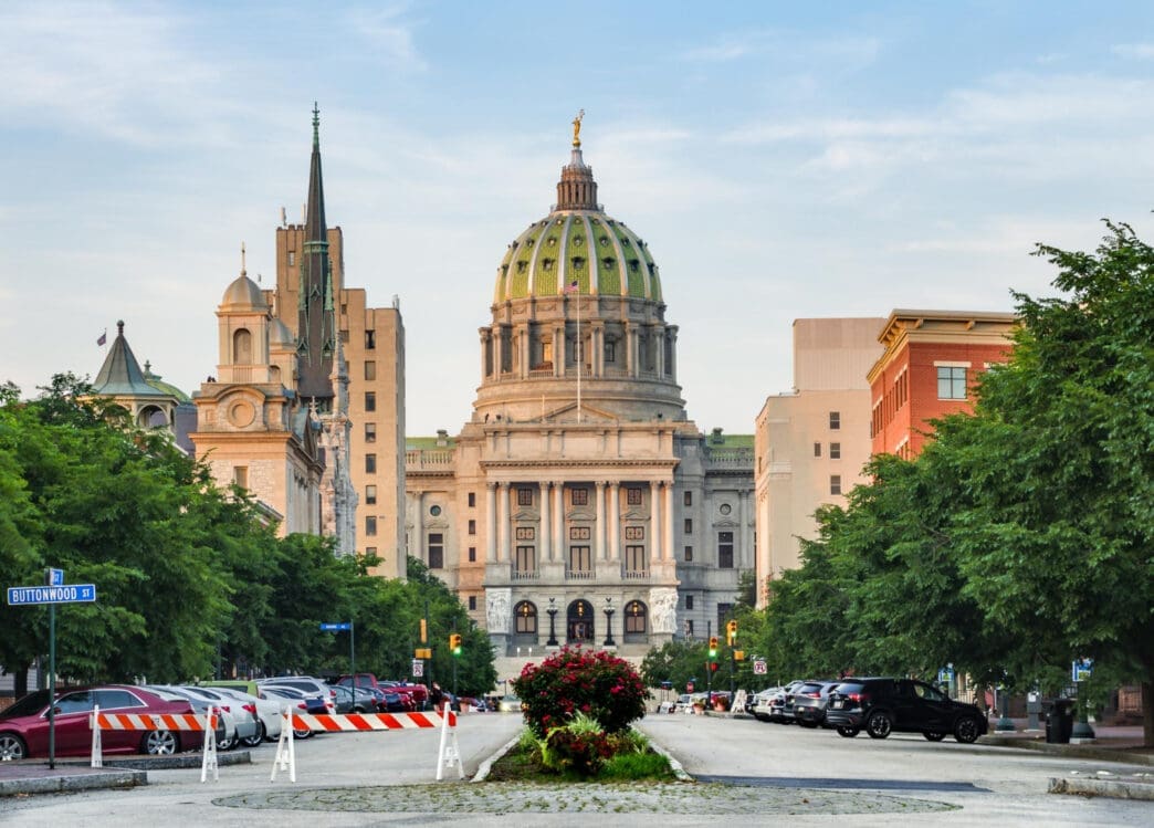 A photograph showing the Pennsylvania State Capitol building with its green dome from the perspective of a tree-lined street with parked cars.