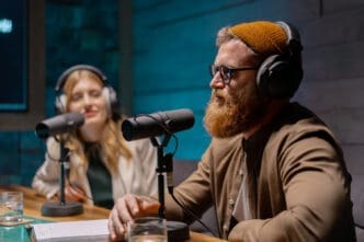 A man with a red beard and a beanie is speaking into a microphone in a podcast studio, with a woman also in headphones and a microphone in the background.