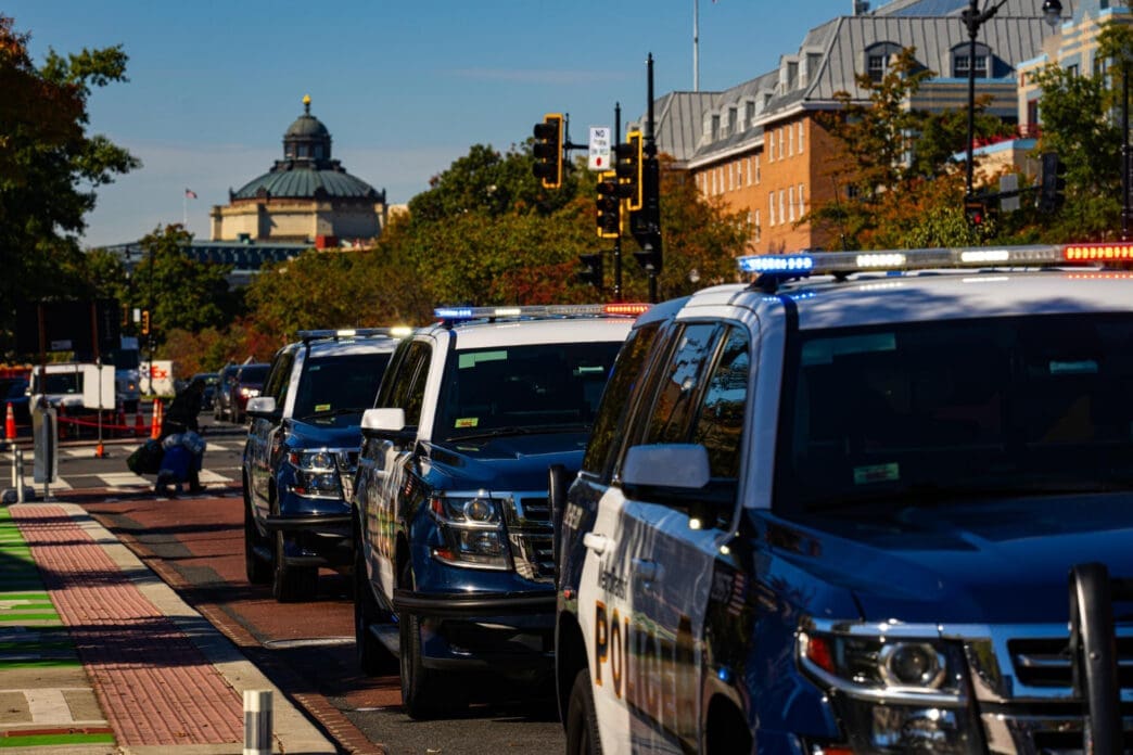 A line of police SUVs with flashing lights are parked on a street in Washington, D.C., with a historic building in the background.