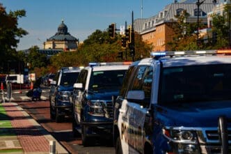 A line of police SUVs with flashing lights are parked on a street in Washington, D.C., with a historic building in the background.
