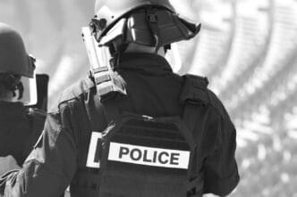 A black and white, close-up shot of a police officer wearing a helmet and a tactical vest with the word "POLICE" on it
