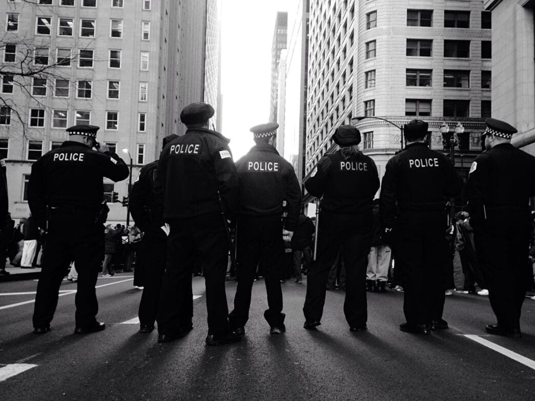 A black and white photograph showing the backs of a line of six police officers standing on a city street between tall buildings, facing a bright light.