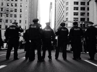 A black and white photograph showing the backs of a line of six police officers standing on a city street between tall buildings, facing a bright light.
