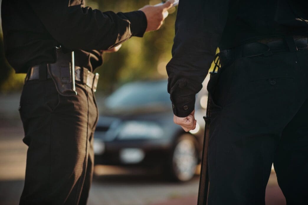 Two police officers stand next to each other, one writing on a piece of paper, with a car in the background
