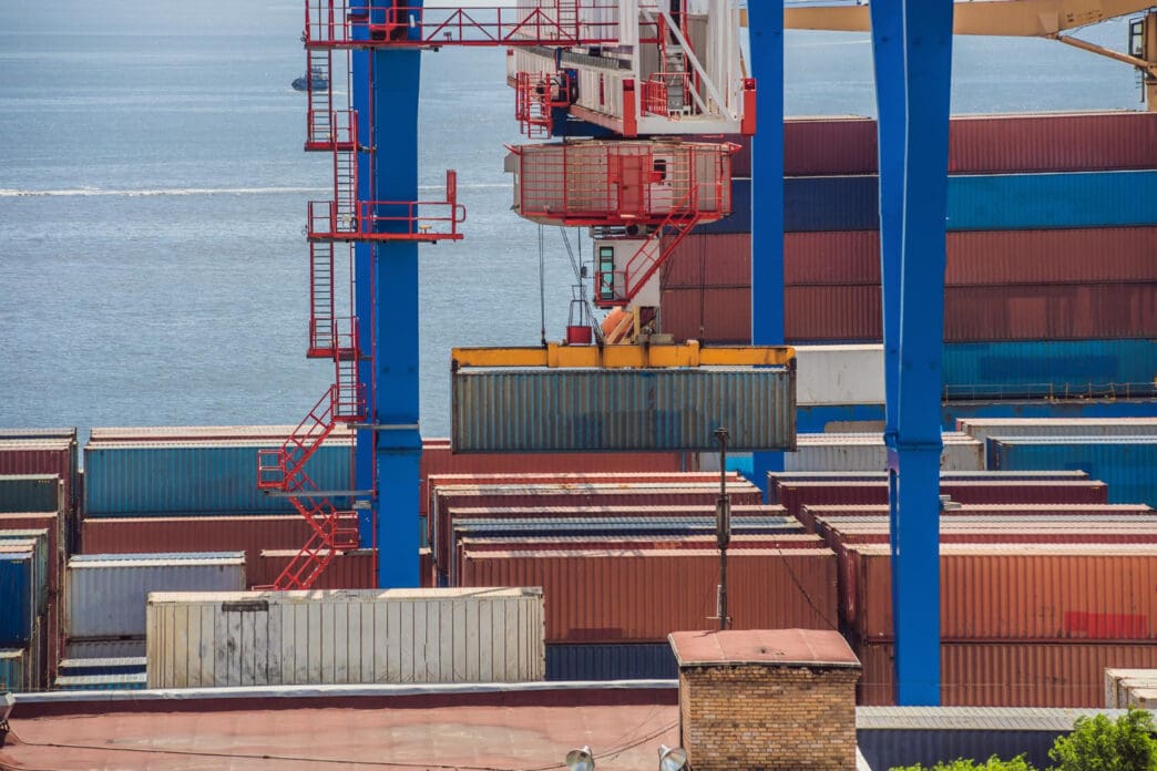 A port crane lifts a large shipping container over a field of stacked containers