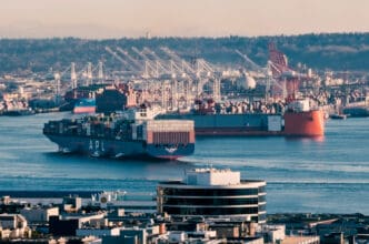 A view of a busy port with two large cargo ships in the water, one labeled 'APL', with numerous cranes and containers visible along the shore in the background.