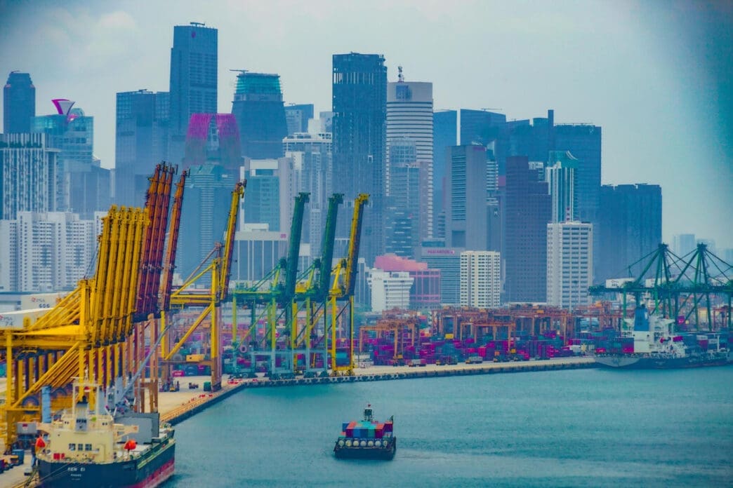 A high-angle view of a bustling port with numerous large cranes, shipping containers, and cargo ships, with the dense, hazy skyline of a modern city in the background.