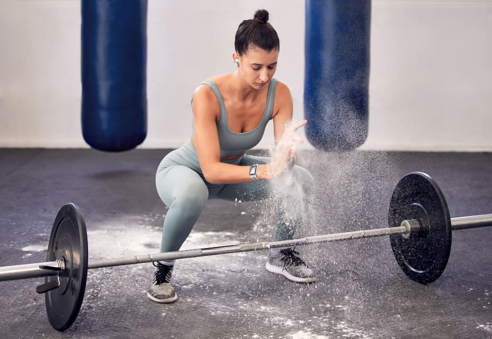 A woman in a gym clapping her hands, creating a cloud of chalk dust, while squatting in front of a barbell.