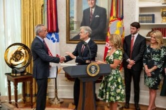 Robert F. Kennedy Jr. shakes hands with Mehmet Oz at a podium during a swearing-in ceremony in the Oval Office, with family members and the large FIFA trophy in the background.