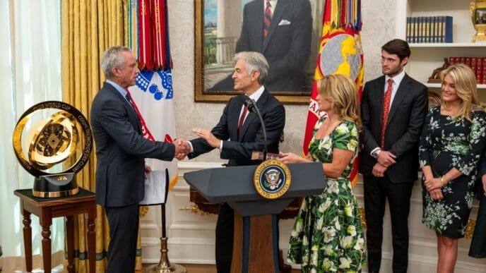Robert F. Kennedy Jr. shakes hands with Mehmet Oz at a podium during a swearing-in ceremony in the Oval Office, with family members and the large FIFA trophy in the background.