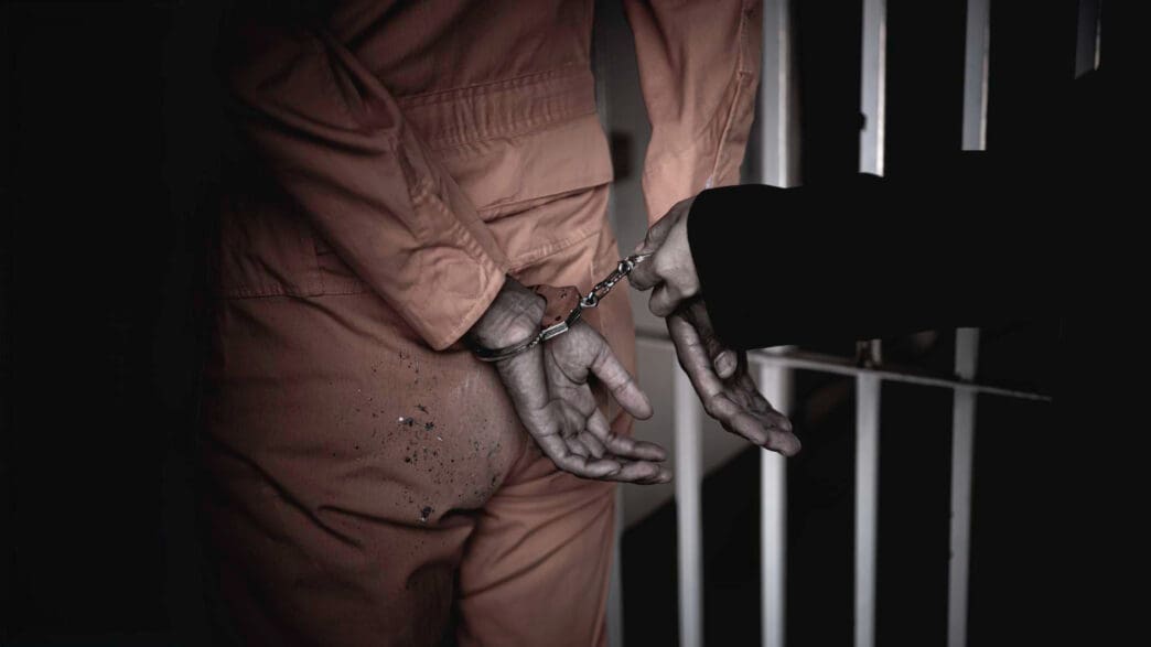 A person in an orange prison jumpsuit is being led by a guard while in handcuffs