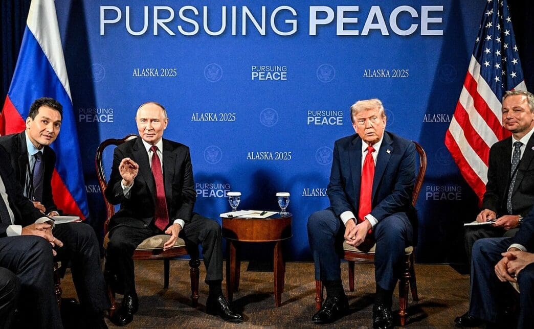 U.S. President Trump and Russian President Putin sit at a conference table with their delegations at the Alaska summit, with flags and a banner behind them.