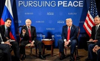 U.S. President Trump and Russian President Putin sit at a conference table with their delegations at the Alaska summit, with flags and a banner behind them.