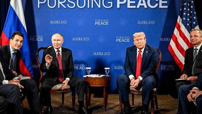 U.S. President Trump and Russian President Putin sit at a conference table with their delegations at the Alaska summit, with flags and a banner behind them.