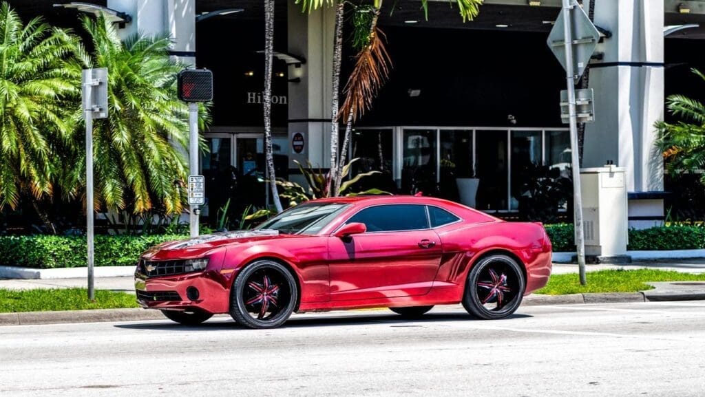 A side-view shot of a shiny, custom red Chevrolet Camaro sports car on a city street in Miami Beach, with a hotel and palm trees in the background.