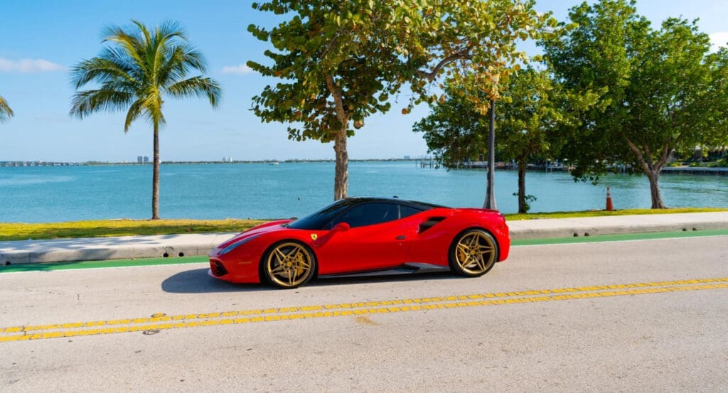 A red Ferrari SF90 Stradale sports car with a black roof and gold rims is parked on a street by a blue waterfront with palm trees.
