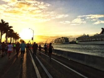 A group of runners is silhouetted against a beautiful sunrise, running along a coastal road lined with palm trees. Two large cruise ships are docked in the background at a port.