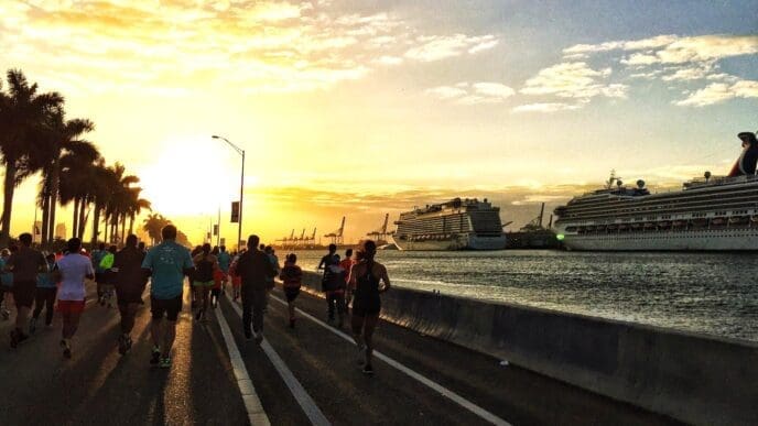 A group of runners is silhouetted against a beautiful sunrise, running along a coastal road lined with palm trees. Two large cruise ships are docked in the background at a port.