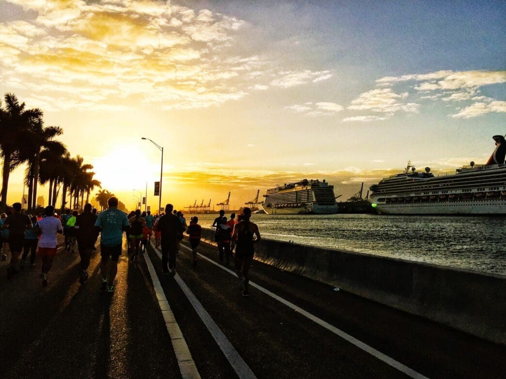 Silhouetted runners and palm trees are visible against a bright sunset with cruise ships in the background.