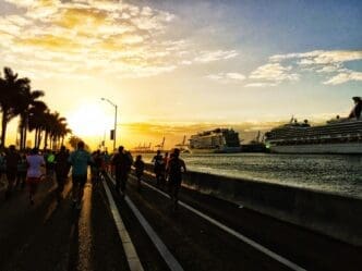 Silhouetted runners and palm trees are visible against a bright sunset with cruise ships in the background.