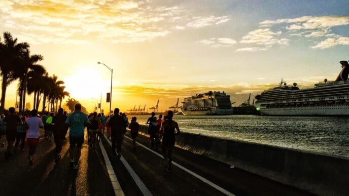 Silhouetted runners and palm trees are visible against a bright sunset with cruise ships in the background.