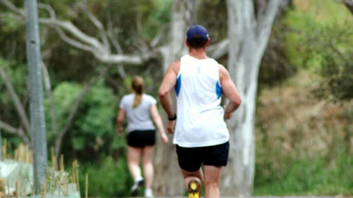 A man runs on a dirt trail in a park, with a woman walking in the background.