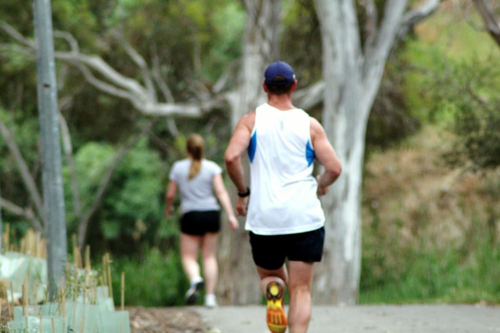 A man runs on a dirt trail in a park, with a woman walking in the background.