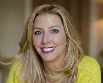 A professional headshot of a smiling Sara Blakely with long blonde hair, wearing a bright yellow sweater, looking directly at the camera.