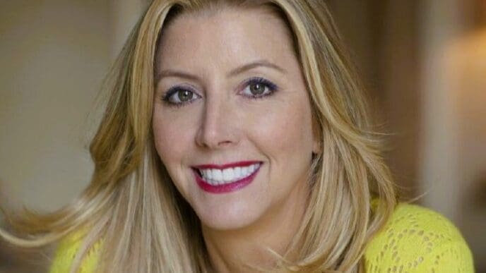 A professional headshot of a smiling Sara Blakely with long blonde hair, wearing a bright yellow sweater, looking directly at the camera.