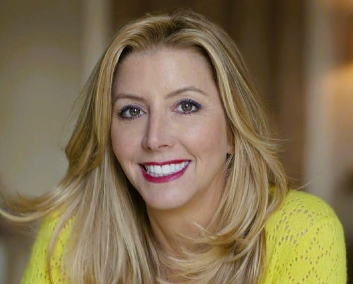 A professional headshot of a smiling Sara Blakely with long blonde hair, wearing a bright yellow sweater, looking directly at the camera.