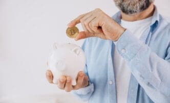 A close-up of a person's hand holding a golden Bitcoin coin over the slot of a white ceramic piggy bank.