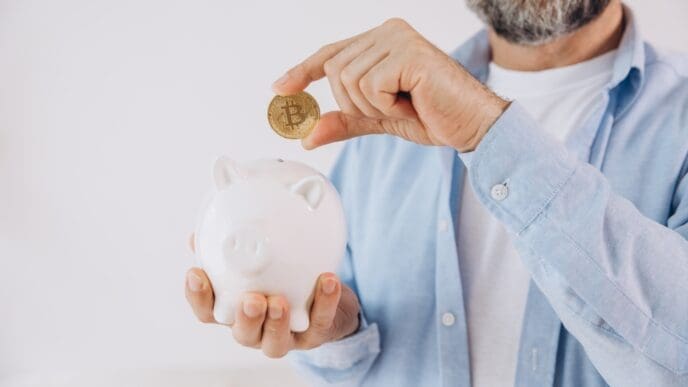 A close-up of a person's hand holding a golden Bitcoin coin over the slot of a white ceramic piggy bank.