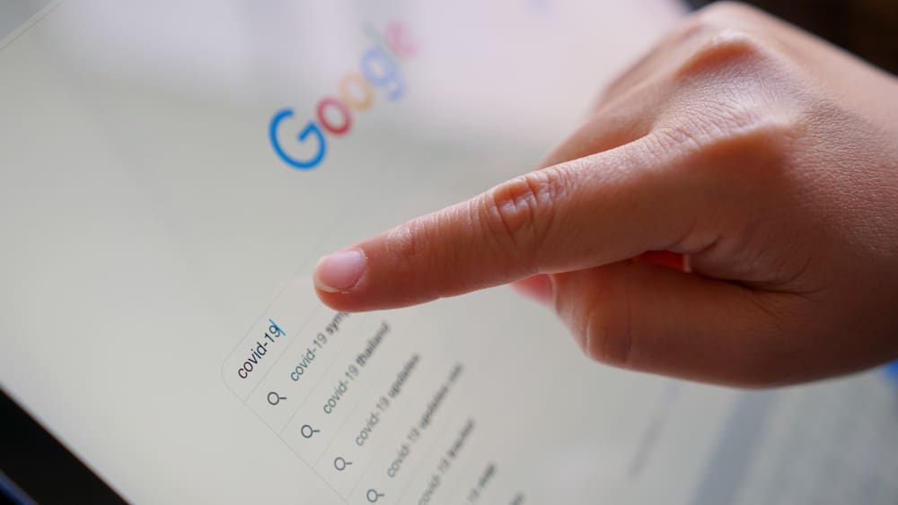 A finger points at a tablet screen displaying the Google search page, with a search query for "covid-19" typed into the search bar and related search suggestions below.