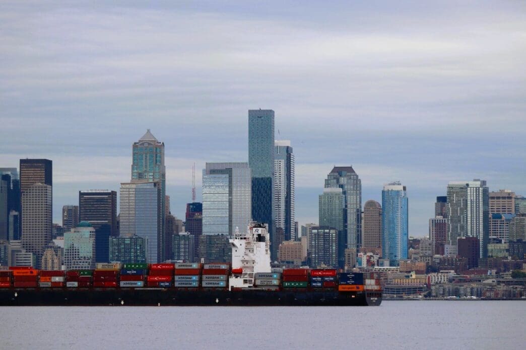 A large cargo ship carrying shipping containers is visible in front of the Seattle skyline