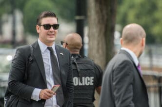 A Secret Service agent in a suit and sunglasses walks in Washington, D.C., with a police officer in the blurred background.