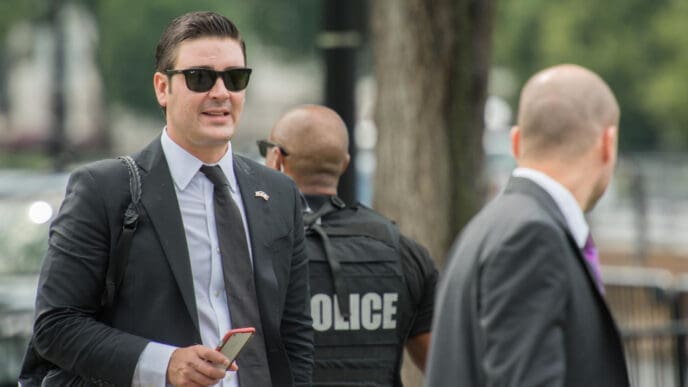 A Secret Service agent in a suit and sunglasses walks in Washington, D.C., with a police officer in the blurred background.