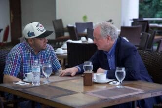 Senator Chris Van Hollen, an older man with gray hair, sits at an outdoor table and talks with a younger man, Kilmar Abrego Garcia, who is wearing a plaid shirt and a baseball cap.