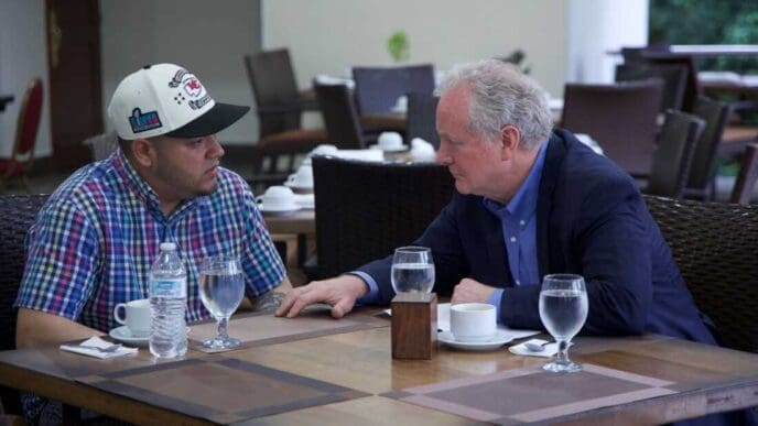 Senator Chris Van Hollen, an older man with gray hair, sits at an outdoor table and talks with a younger man, Kilmar Abrego Garcia, who is wearing a plaid shirt and a baseball cap.