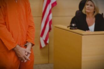 A blurry image of a person in an orange jumpsuit in a courtroom, with a judge visible in the background.
