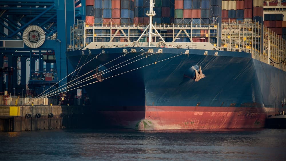 A close-up, frontal view of a large blue container ship with a red hull, docked at a port and loaded with multi-colored shipping containers.